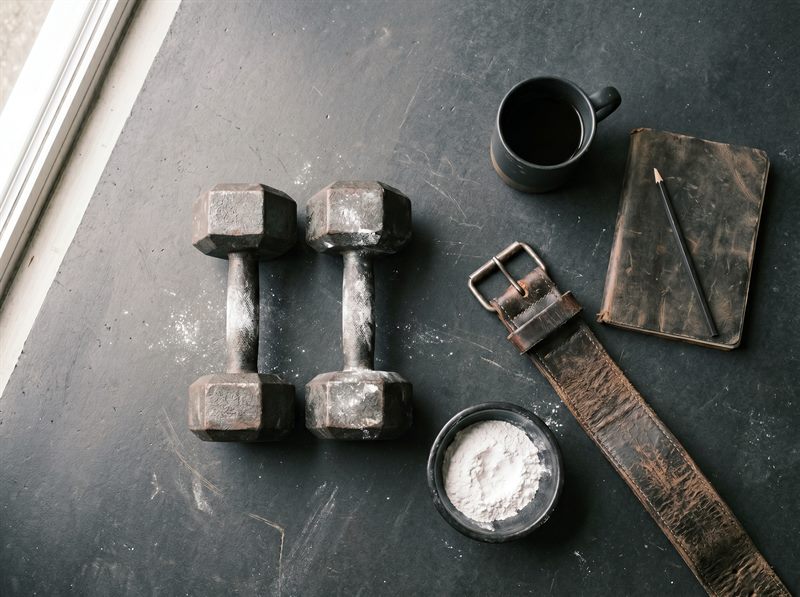 A lifter's kit on dark concrete: worn dumbbells dusted with chalk, a leather belt, chalk, a notebook and coffee.