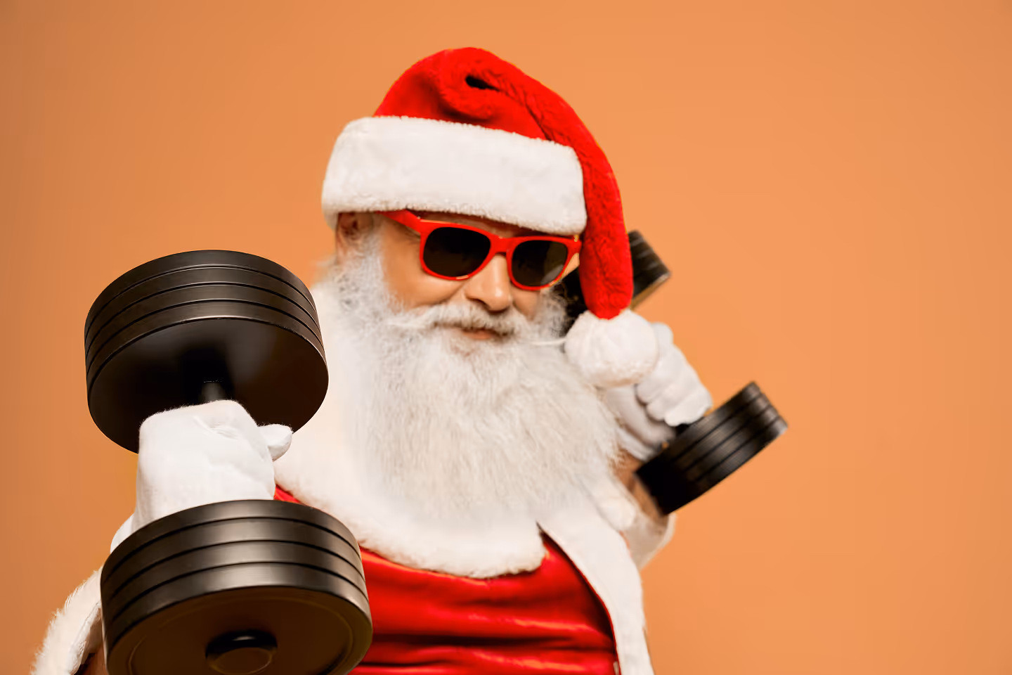 Santa Claus wearing red sunglasses and hat, lifting two black dumbbells against a plain mustard background.