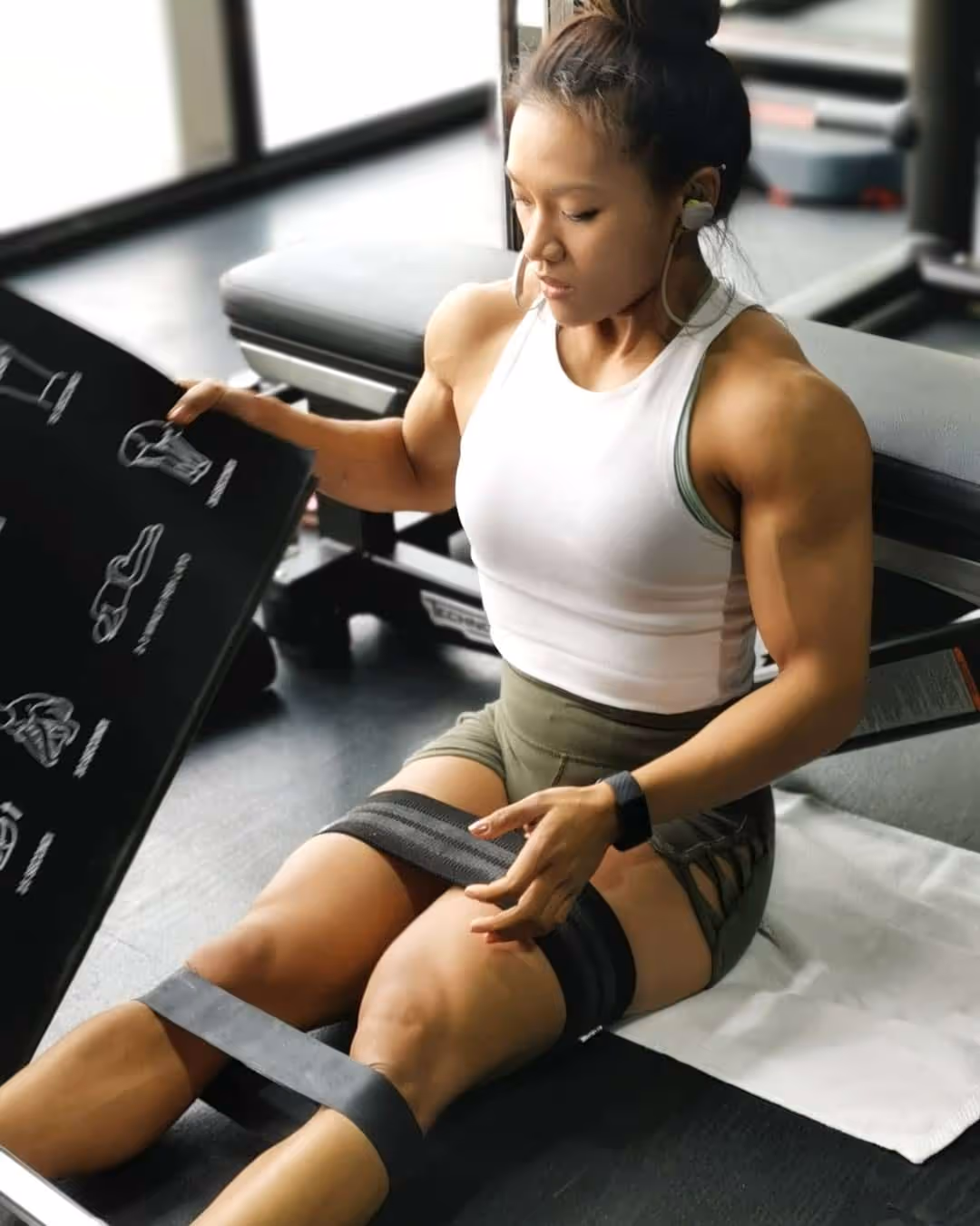 Muscular woman in a white tank top and green shorts sits on the gym floor wrapping resistance bands around her thighs and legs.