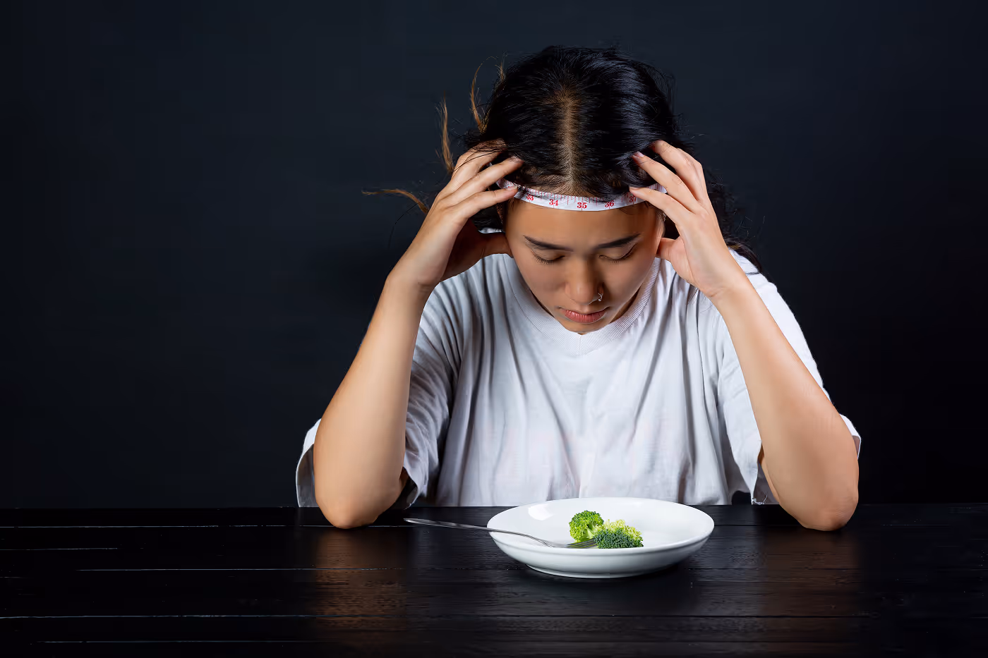 Woman with a measuring tape around her forehead looks down at a white plate with two small broccoli florets, appearing distressed.