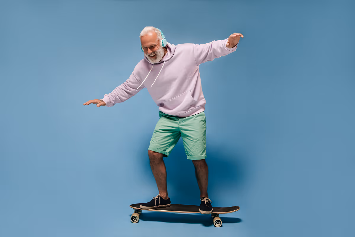 Smiling elderly man in a lavender hoodie and green shorts balancing on a skateboard against a blue background while wearing headphones.