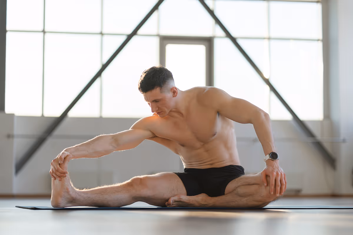 Shirtless man in black shorts stretching seated on a yoga mat in a bright gym.